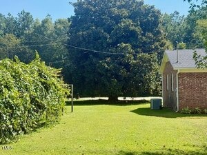 A grassy backyard with a brick house on the right, a large tree in the background, a hedge on the left, and overhead utility lines crossing the yard. An air conditioning unit is next to the house.