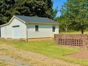 A single-story white garage with a gray roof stands on a grassy lawn beside a small brick wall with decorative openings. Trees and a clear sky are visible in the background.