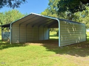 A metal carport with an open front and sides and a covered roof sits on a grassy area, with trees and another small structure visible in the background. Sunlight casts shadows on the ground.