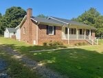 Charming single-story brick house at 6527 US 15, Oxford, featuring a covered front porch with white railing and gabled roof. Surrounded by lush grass and trees, it also includes a small outbuilding and a concrete path to the porch steps.