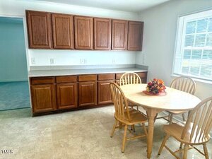 A kitchen with wooden cabinets, a light-colored countertop, and a round wooden table with four matching chairs. A decorative centerpiece is on the table, and there is a window letting in natural light.