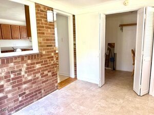 A sunlit indoor area with brick and white walls, tile flooring, a small closet with folding doors, and an open window counter looking into a kitchen with wooden cabinets. A broom and dustpan are visible in the closet.