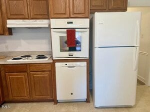 A white refrigerator stands next to a built-in white oven with a red towel hanging on its handle, a white dishwasher below, and a white stove with a range hood, all surrounded by brown wooden cabinets.