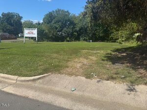 A grassy vacant lot bordered by a sidewalk and street, with a white real estate sign in the background and trees along the edges under a clear blue sky.