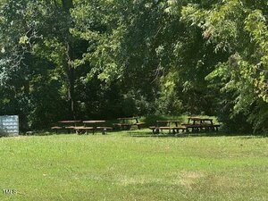 Several wooden picnic tables are arranged in groups on a grassy area surrounded by trees and dense greenery. Sunlight filters through the leaves, casting dappled shadows on the ground.