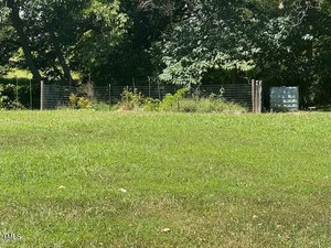 A grassy lawn leads up to a fenced garden area with various plants, surrounded by trees and partial shade. A metal structure or container is visible to the right of the fenced area.