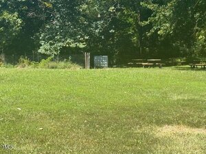 A grassy field with a few picnic tables near the edge, bordered by a wire fence and trees in the background. Sunlight filters through the foliage, casting patches of light on the grass.