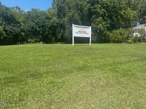 A grassy lawn with a white sign that reads "Future Home of The Hope House" stands in the center; trees and shrubs line the background under a clear blue sky.