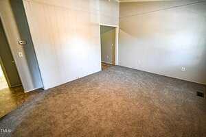 A room with light gray paneled walls, gray carpet flooring, an open doorway leading to another area, and a small section of tile flooring visible near the entrance. Electrical outlets are on the walls.