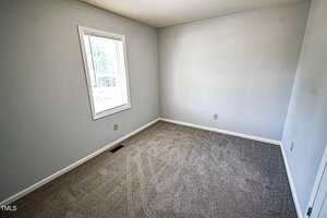 A small empty room with light gray walls, a window with white blinds, beige carpet flooring, and a white ceiling. Electrical outlets and an air vent are visible. Sunlight comes through the window.