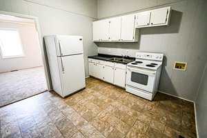 A small kitchen with white cabinets, a white refrigerator, a white electric stove, a stainless steel sink, and tile flooring. An open doorway leads to a carpeted room with a window.