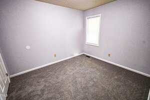 A small, empty room with light gray walls, a single window with closed blinds, a white door, and gray carpeted flooring. There are several electrical outlets visible on the walls.