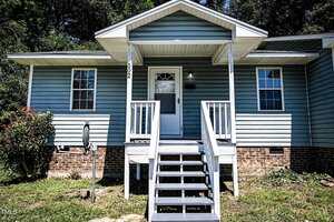 A blue single-story house with white trim, a small covered front porch, white railings and steps, brick foundation, two windows, and a grassy front yard with a bush on the left side.
