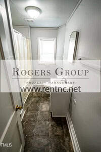 A narrow bathroom with gray tile flooring, a small white vanity with a mirror above it, a window with blinds, and a shower curtain on the left. The Rogers Group property management watermark is over the image.