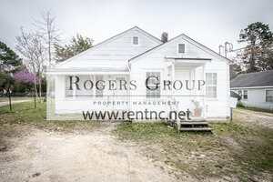 A small, white, single-story house with a front porch and two front doors, situated on a patchy grass and dirt yard. The house has simple exterior features and a few potted plants near the steps.