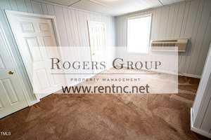 A small, empty room with carpeted floors, light gray wood-paneled walls, two white doors, a window, and a wall-mounted air conditioning unit. The Rogers Group logo and website are overlaid on the image.
