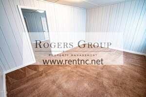 Empty room with beige carpet, white paneled walls, and a white ceiling. There is an open doorway leading to another room. A logo for Rogers Group Property Management and a website overlay the image.