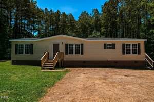A single-story manufactured home at 141 Cardinal Lane, Henderson, with beige siding, black shutters, front steps with railings, surrounded by trees and sitting on a grassy lawn with a dirt driveway in front.