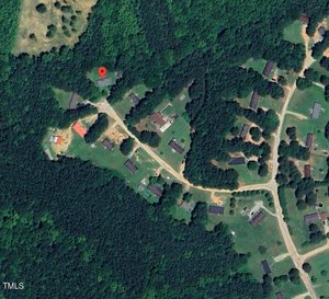 A satellite view of a rural residential area with scattered houses, winding roads, open green spaces, and dense surrounding forests. A location marker pinpoints one of the houses near the tree line.