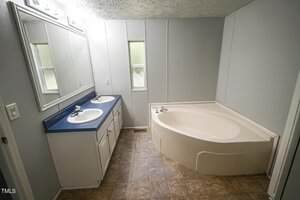 A bathroom with two sinks set in a blue countertop on a white vanity, a large corner bathtub, light-colored walls, a large mirror, and two narrow frosted windows above the sinks.