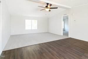 A bright, empty room with light-colored walls, a ceiling fan with lights, a large window, carpeted flooring on one side, and dark wood flooring on the other. An open doorway leads to another room.