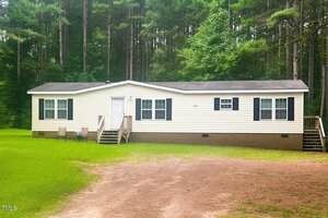 A beige manufactured home with black shutters sits on a brick foundation surrounded by grass and trees. Two sets of steps lead to entrances, and a small table with two chairs is near the front door.