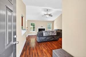 A living room with tan walls, wood flooring, two dark sofas, a fireplace, ceiling fan, and large windows letting in natural light. The image is taken from the open front door.