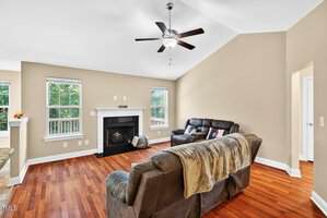 A living room with wood floors, beige walls, two brown sofas, a fireplace with a white mantel, a ceiling fan with lights, and two windows showing greenery outside. A doorway is visible on the right.