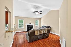 A living room with beige walls, wood flooring, a ceiling fan, a fireplace between two windows, and two dark sofas facing the fireplace. A granite counter is visible on the left.