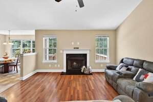 A living room with beige walls, two windows, a dark fireplace with logs and baskets, and a gray sectional sofa. The hardwood floor leads to a partially visible dining area with tile flooring and a hanging light fixture.