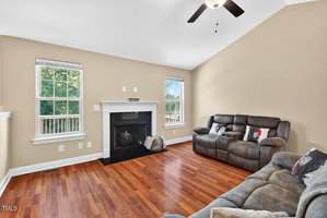 A living room with tan walls, hardwood floors, two gray reclining sofas, a fireplace with a white mantel, and two windows letting in natural light. A ceiling fan is mounted on the vaulted ceiling.