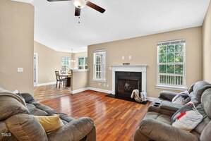 A living room with hardwood floors, beige walls, two gray sofas, a ceiling fan, and a fireplace. Large windows let in natural light. The dining area is visible in the background.