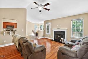 Living room with hardwood floors, beige walls, a ceiling fan, and a black fireplace. Two recliners face the fireplace. Open view to a kitchen with wood cabinets and a breakfast bar. Large windows let in natural light.