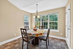 A small dining area with three wooden chairs around a round table covered by a sunflower-patterned tablecloth. Large windows let in natural light and provide a view of greenery outside. A ceiling light hangs above the table.