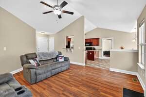 A living room with wood flooring, gray reclining sofa, ceiling fan, and beige walls. The area opens to a kitchen with tile flooring and dark wood cabinets. Natural light enters through a window on the right.