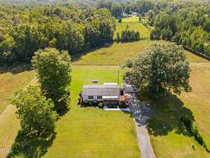 Aerial view of a single-story house with a gray roof at 6688 Goshen Road, Oxford, surrounded by a large, well-maintained lawn, trees, and open fields, with a driveway and wooded area in the background.