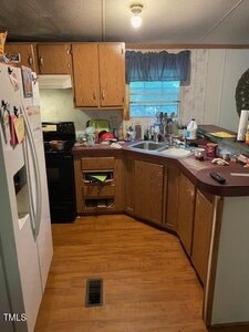 A small kitchen with wood cabinets, a white fridge, a black stove, a double sink, and a cluttered countertop with dishes, cans, and cleaning supplies. The floor is wood, and there is a window with a blue valance.