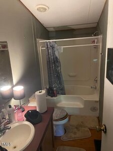 A small bathroom with a sink and counter on the left, a toilet in the center, and a bathtub-shower combo with a curtain at the back. Various toiletries are on the counter and the edge of the tub.