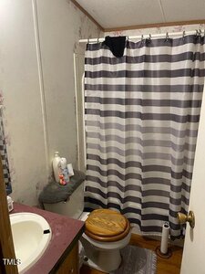 A small bathroom with a sink, a brown toilet seat, toiletries on the tank, a gray and white striped shower curtain, and a black towel draped over the curtain rod.