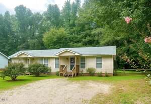 Charming single-story beige house at 228 Crozier Street, Henderson, with a gray roof, small porch with steps, and several windows. Gravel driveway and bushes in front. Enjoy a grassy yard with dense green trees in the background.