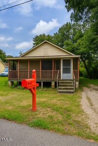 A small, single-story house with a screened front porch and steps leading to the entrance. A bright red mailbox is positioned near the street, and trees surround the property.