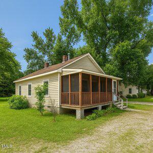 This single-story beige house with white trim and a screened front porch sits on a grassy lawn with large trees and a gravel driveway at 436 John Street in Henderson.
