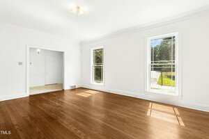 A bright, empty room with hardwood floors, white walls, two tall windows letting in natural light, and an open doorway leading to another room with tile flooring.
