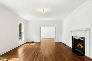 A spacious, empty living room with white walls, hardwood floors, two large windows, a lit fireplace, and an open doorway leading to another bright room.