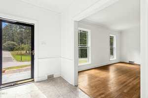 A view of a home's interior shows a tiled entryway with an open door and adjacent room featuring hardwood floors, white walls, and large windows overlooking a green yard.