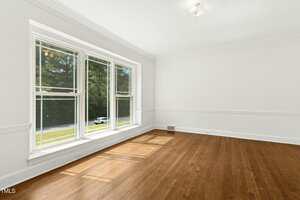 Empty room with white walls, a ceiling light, and hardwood flooring. Large windows on one wall let in natural light, with a view of trees and a road outside.