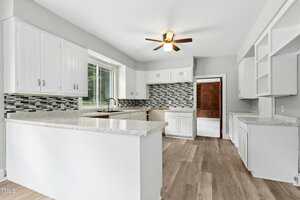 Bright kitchen with white cabinets, light countertops, a large window over the sink, gray walls, wood-style flooring, a ceiling fan, and a gray and white mosaic tile backsplash. Open doorway leads to another room.