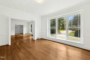 A spacious, empty room with hardwood floors, white walls, and large windows letting in natural light. Another open room is visible through a wide doorway, and a street with greenery can be seen outside.