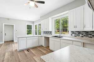 Bright kitchen with white cabinets, marble countertops, a double sink under large windows, a gray and black tile backsplash, light wood floors, and a ceiling fan. Door and thermostat are visible in the background.