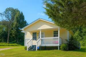 A small, yellow, single-story house with white trim and a front porch with railings sits on a grassy lawn surrounded by trees on a sunny day. A front door and two windows face the porch steps.
